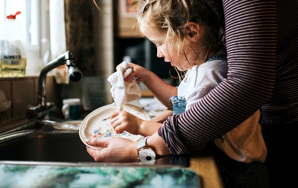 A girl is seen cleaning the dishes by the sink. Someone is helping her but the face is not seen.