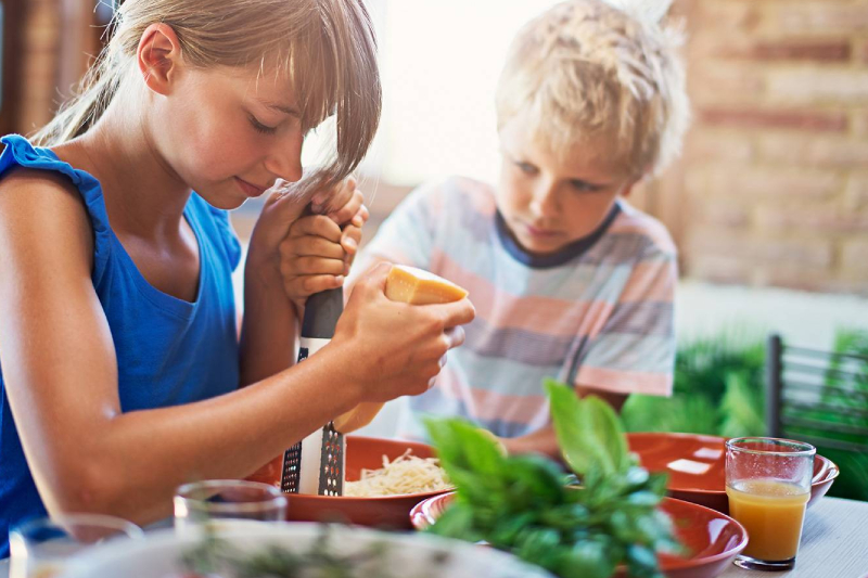 two children are in the picture. One of them, a girl, is grating cheese, while the other, a boy is watching her.